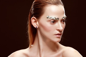 Close-up of gold eyebrow jewelry and blue gemstone studs worn by a model in soft light