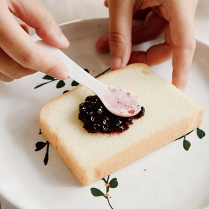 Natural white shell spoon spreading purple blueberry jam on toast in a green floral plate
