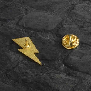 Creative gold lightning bolt enamel pin with butterfly clutch back on grey stone background