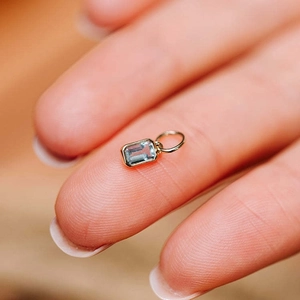 Gold stainless steel birthstone charm with light blue rectangular zircon on fingertip