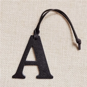 Close-up of black lychee leather A-letter bag charm on a soft beige linen background
