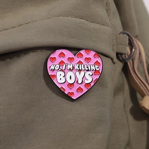 Close-up of pink heart enamel pin with NO, I'M KILLING BOYS slogan on olive canvas