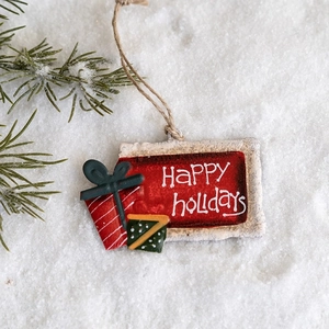 Vintage red iron gift box ornament with Happy Holidays text hanging on a snowy pine tree branch