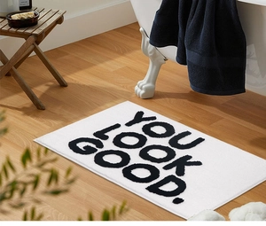 Detail of white jacquard bath mat with black 'YOU LOOK GOOD' text on wood floor