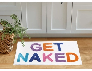 Close-up of white plush bath mat with colorful GET NAKED 3D jacquard text on wood floor