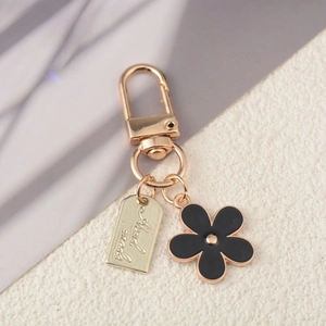 Golden keychain featuring a black enamel flower and a handmade engraved metal tag