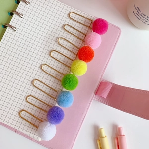 Detailed view of eight colorful pompom paper clips clipped to a pink grid notebook