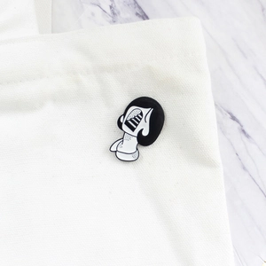 Surrealist enamel skull mask pin displayed on a white canvas fabric bag surface