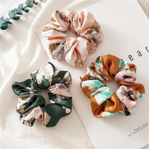 Three satin floral scrunchies in pink, dark green, and orange on a lifestyle background