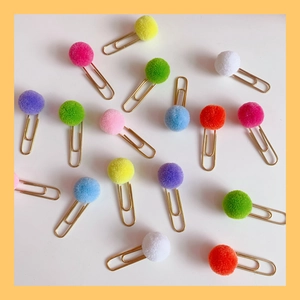 Flat lay of golden paper clips with colorful fluffy pom poms on a clean white background