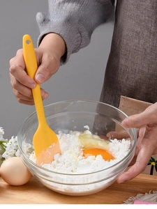Macro view of small yellow silicone spatula mixing eggs and flour in a glass bowl