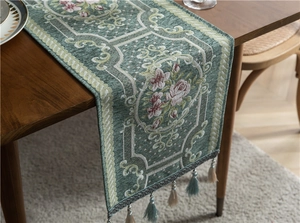 Detail of teal chenille table runner with pink floral embroidery and tassels on wood table