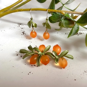 Close-up of orange fruit brooch and earrings with metal leaves on white texture