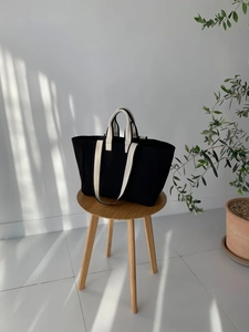 Minimalist black canvas tote bag with beige straps on a wooden stool in soft natural light