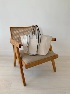 Detail view of a cream canvas tote bag with black piping handles on a wooden chair