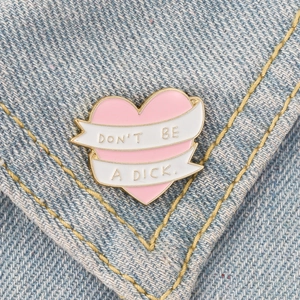 Close-up of pink heart enamel pin with 'Don't Be A Dick' text on a denim jacket
