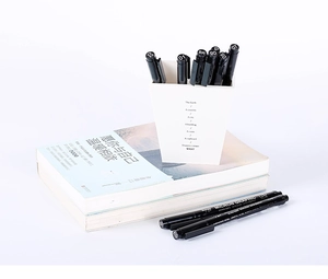 Waterproof drawing pens with various nib sizes arranged on a book next to a white pen holder