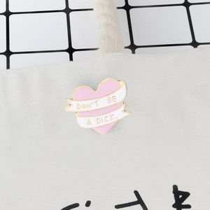Pink heart enamel brooch with 'Don't be a dick' ribbon on canvas bag surface