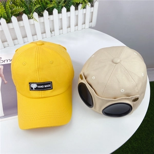 Yellow baseball cap and beige aviator hat with signature goggles on a white table