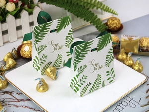 Close-up of white paper favor box with green fern patterns and gold Save the Date text
