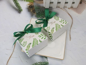 Detail of square paper favor box with green leaf patterns and silk ribbon on a plate