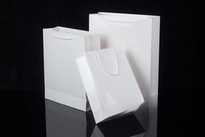 Three sizes of thick white gift bags with rope handles on a black reflective surface