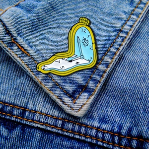Detail of melting clock enamel pin with yellow border on blue denim jacket fabric