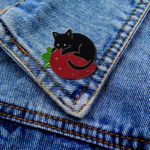 Detail of a cartoon black cat on a red strawberry enamel pin pinned on blue denim fabric