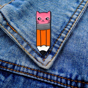 Detail of pink cat and orange pencil enamel pin on blue denim jacket fabric