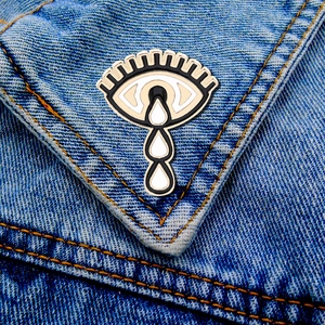 Detail of teary eye enamel pin on blue denim fabric background with gold metal edges