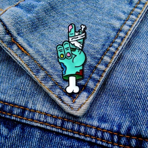 Close-up of zombie hand enamel pin with V sign and bandages on blue denim jacket