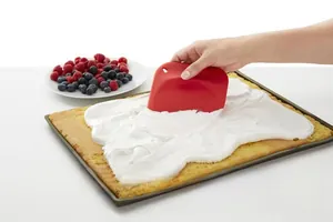 Red silicone baking spatula spreading white cream on a square sponge cake base