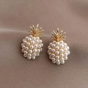 Close-up of golden pineapple stud earrings with white pearls on a soft fabric background