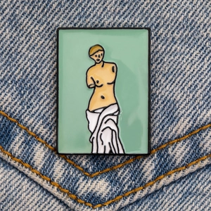 Détail du pin's émaillé Vénus de Milo sur denim bleu avec un fond vert artistique