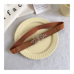 Detail of a brown elastic belt featuring a golden metallic leaf buckle on beige linen
