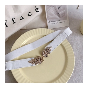 Detail of white elastic waist belt featuring golden leaf-shaped metal buckle on a plate