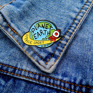 Cartoon Planet Earth Space Cadet enamel pin on a blue denim jacket pocket