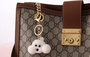Close-up of a white plush puppy keychain hanging on a brown bag with gold hardware