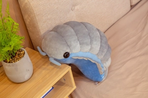 Detail view of large grey and blue plush woodlouse pillow on a cozy bed with plant
