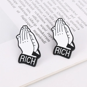 Détail des broches émaillées RICH noir et blanc avec mains en prière sur un livre ancien