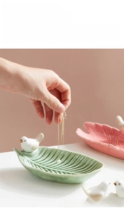Detail showing a hand placing jewelry into a leaf-shaped ceramic tray with a bird figurine
