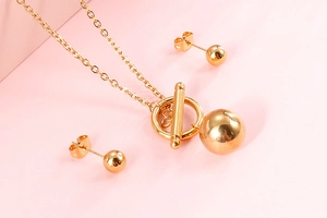 Detail of gold OT toggle necklace and matching spherical stud earrings set on pink background