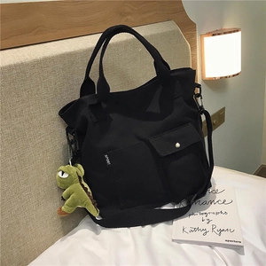 Detail of black canvas tote bag with green dinosaur plush charm on a cozy white bed