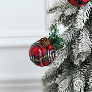 Plaid Christmas ball ornament with red fabric, pine cones and needles hanging on a snowy tree