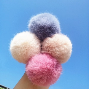 Fuffy faux fur pompom hair ties in purple, beige and pink against a clear blue sky background