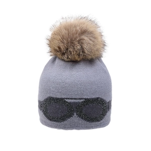 Detailed close-up of grey knit beanie with large faux fur pom and decorative goggles