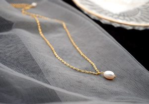 Detailed view of a gold chain necklace featuring a white teardrop pearl on soft grey fabric