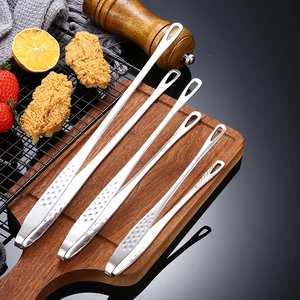 Five-piece stainless steel BBQ tongs set on a rustic wooden board with kitchen props