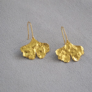Macro shot of gold-plated ginkgo leaf earrings with textured finish on gray fabric