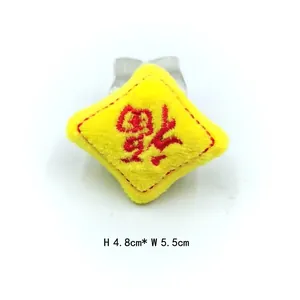 Détail d'un charm en peluche jaune avec broderie rouge et clip en métal, 5,5 cm de large